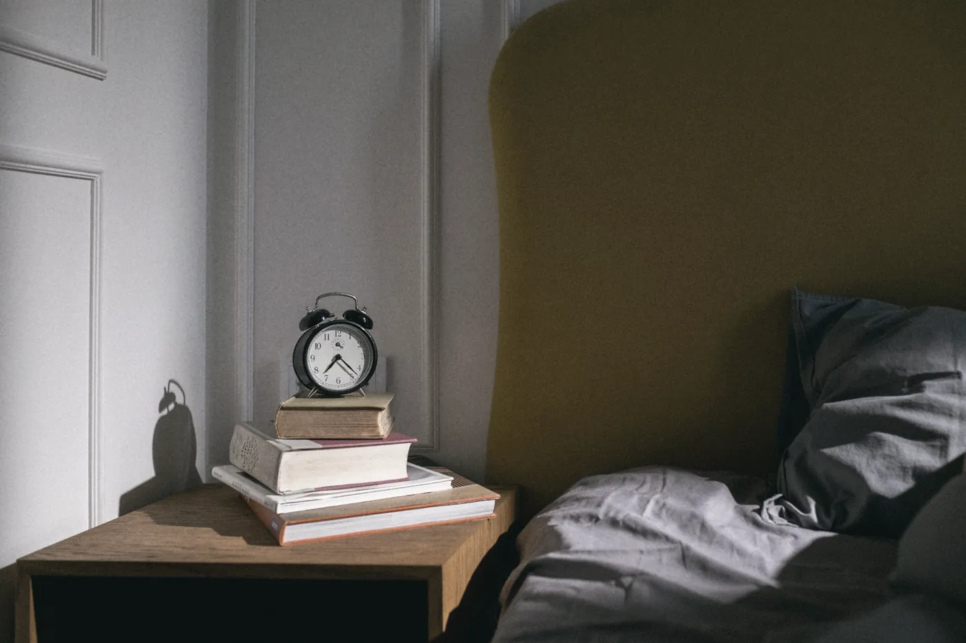 An alarm clock and books on a bedside table beside an empty bed
