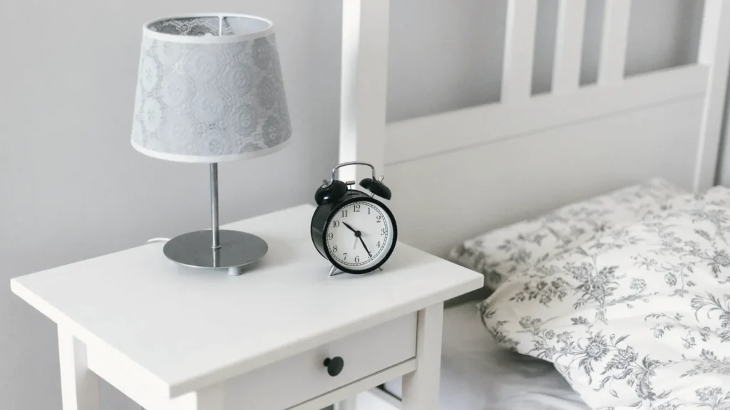 A white bedside table with a lamp and an alarm clock next to a bed