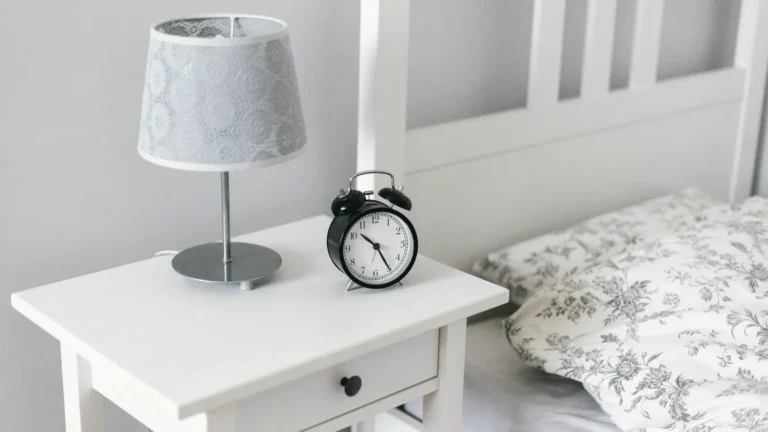 A white bedside table with a lamp and an alarm clock next to a bed